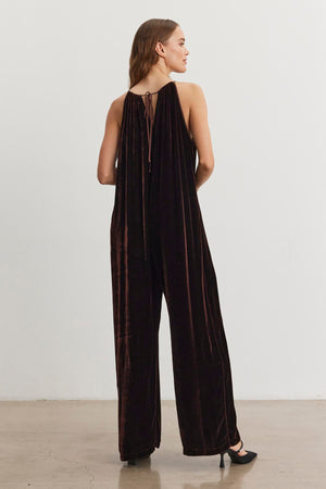 A woman stands with her back to the camera in a neutral room, wearing the Cienna Silk Velvet Jumpsuit by Velvet by Graham & Spencer, paired with black heels.