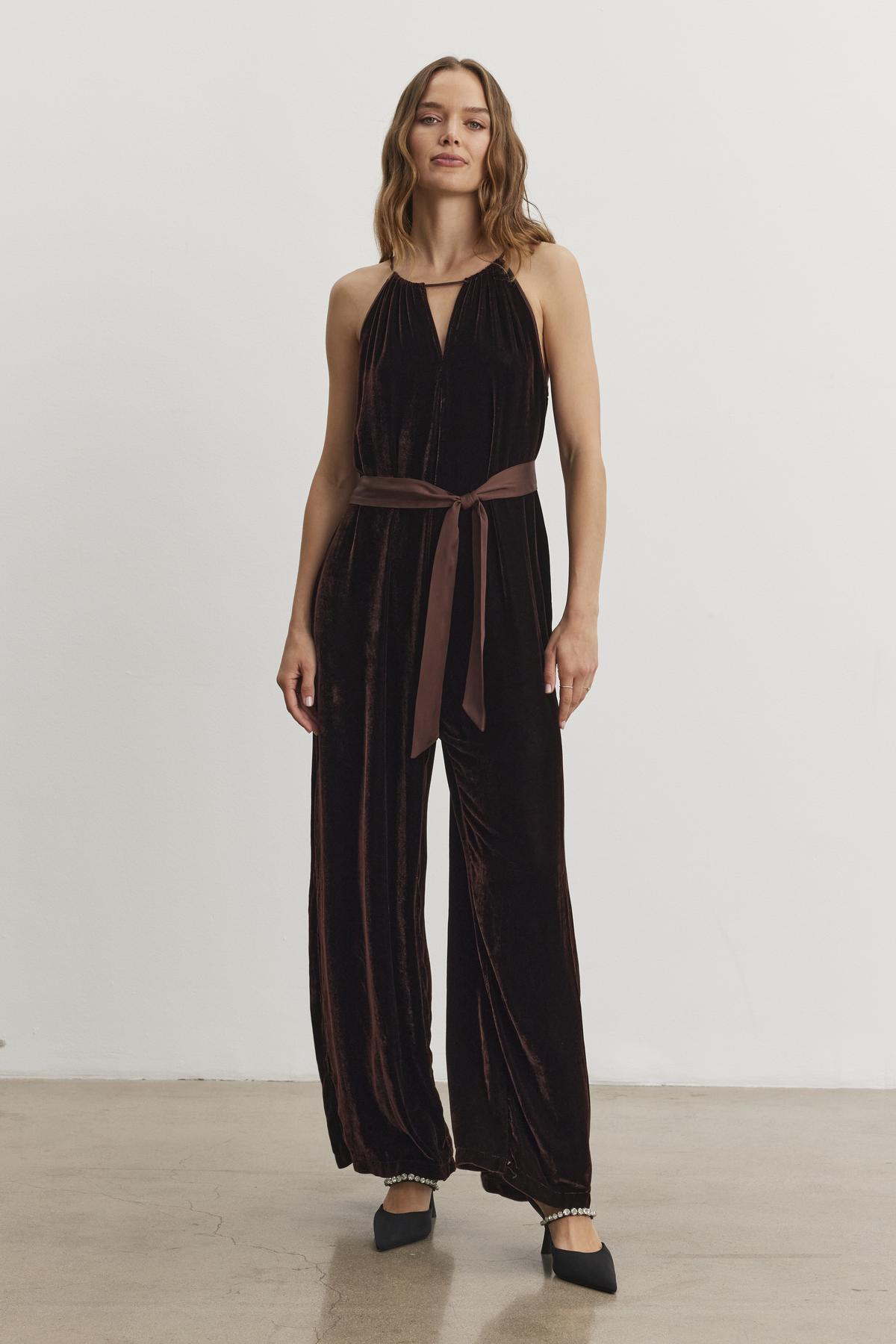 A person is modeling the "CIENNA SILK VELVET JUMPSUIT" by Velvet by Graham & Spencer, featuring a dark color and a tied waist belt, set against a plain backdrop.-38246397378753