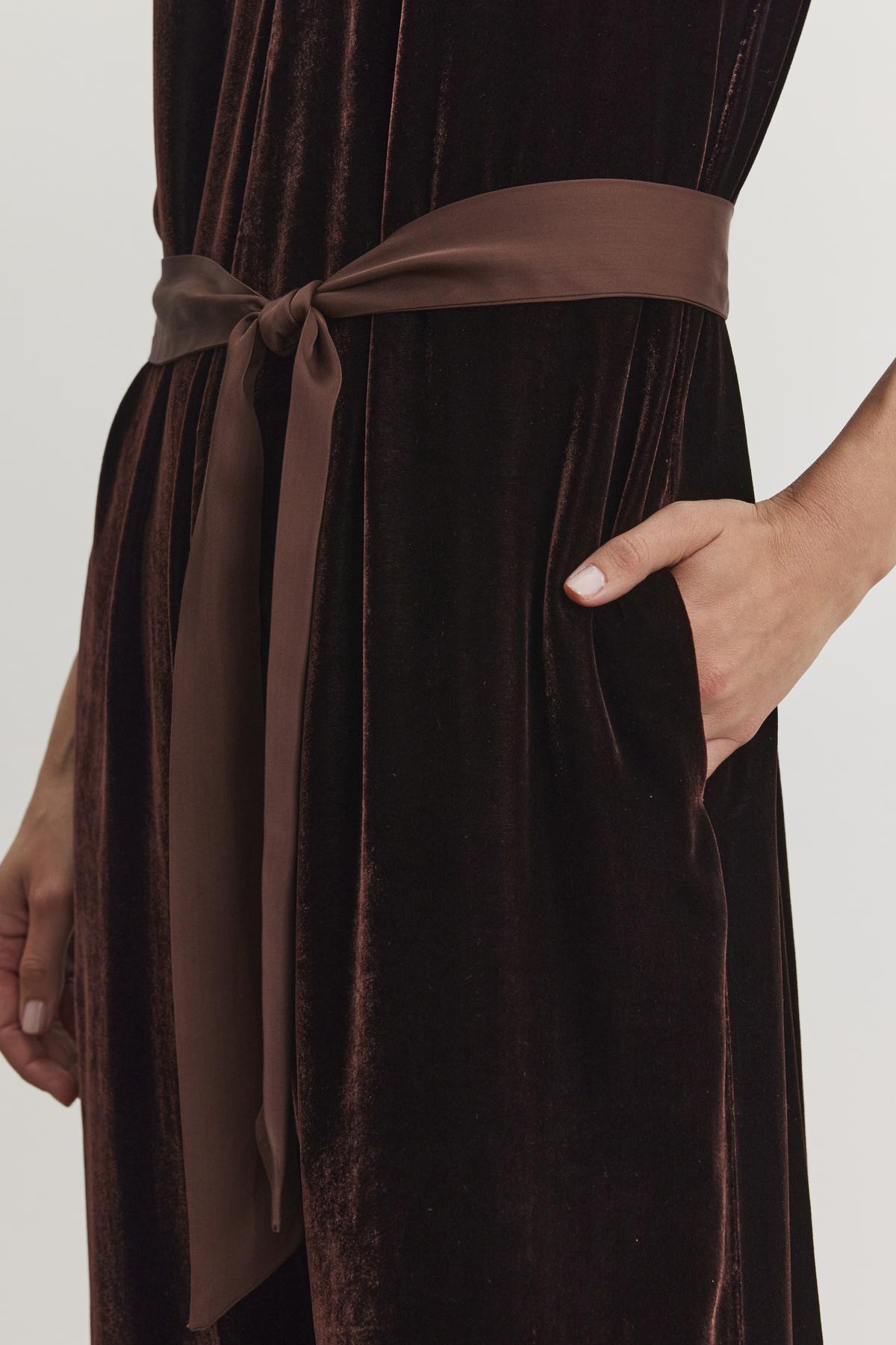 A person wearing the CIENNA SILK VELVET JUMPSUIT from Velvet by Graham & Spencer, featuring a dark hue with a brown satin sash tied at the waist and hands casually placed in pockets.-38246397411521