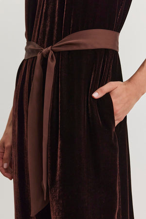 A person wearing the CIENNA SILK VELVET JUMPSUIT from Velvet by Graham & Spencer, featuring a dark hue with a brown satin sash tied at the waist and hands casually placed in pockets.