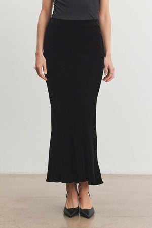 An individual is standing with the EZA Silk Velvet Skirt by Velvet by Graham & Spencer and sleek black pointed shoes on a polished floor, set against a plain white background.