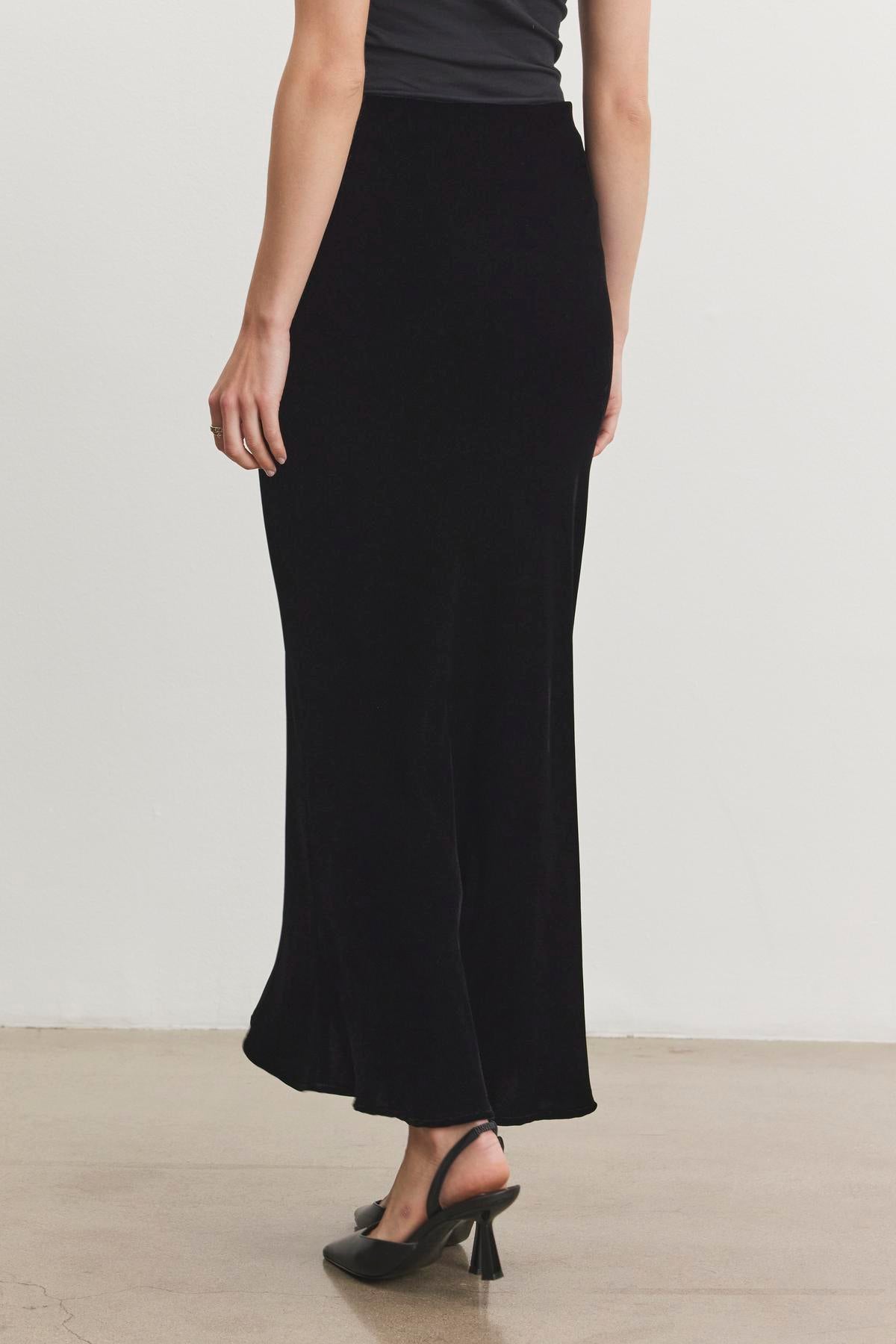 A person wearing the EZA Silk Velvet Skirt by Velvet by Graham & Spencer and a black top stands on a light-colored floor, facing away. They complete the look with black heels.-38246461702337