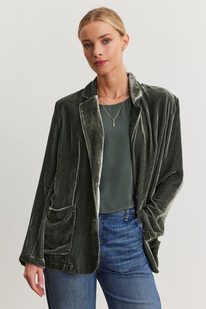 A person wearing the KYLA SILK VELVET BLAZER by Velvet by Graham & Spencer, along with a green top and blue jeans, stands against a plain background.