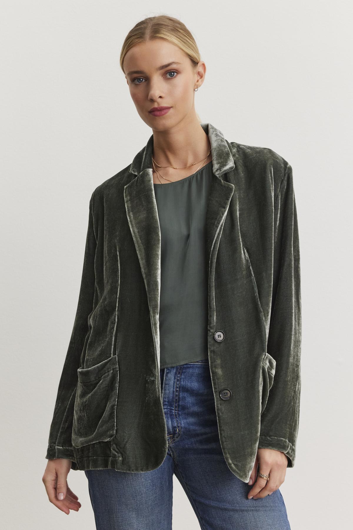 A person wearing the KYLA Silk Velvet Blazer from Velvet by Graham & Spencer, featuring an oversized fit in dark green with a notched collar, and paired with a green top and jeans, stands against a plain white background.