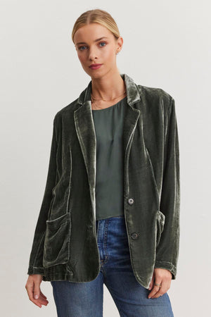 A person wearing the KYLA Silk Velvet Blazer from Velvet by Graham & Spencer, featuring an oversized fit in dark green with a notched collar, and paired with a green top and jeans, stands against a plain white background.