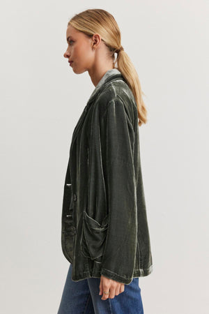 A woman with blonde hair in a ponytail models the KYLA SILK VELVET BLAZER by Velvet by Graham & Spencer, along with blue jeans, standing in profile against a neutral background.