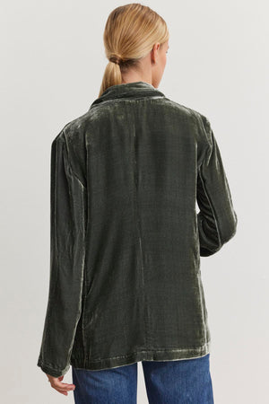 Against a simple backdrop, an individual with blonde hair styled in a ponytail is seen from behind wearing an oversized KYLA Silk Velvet Blazer in dark green, featuring a notched collar. The blazer is from the brand Velvet by Graham & Spencer.