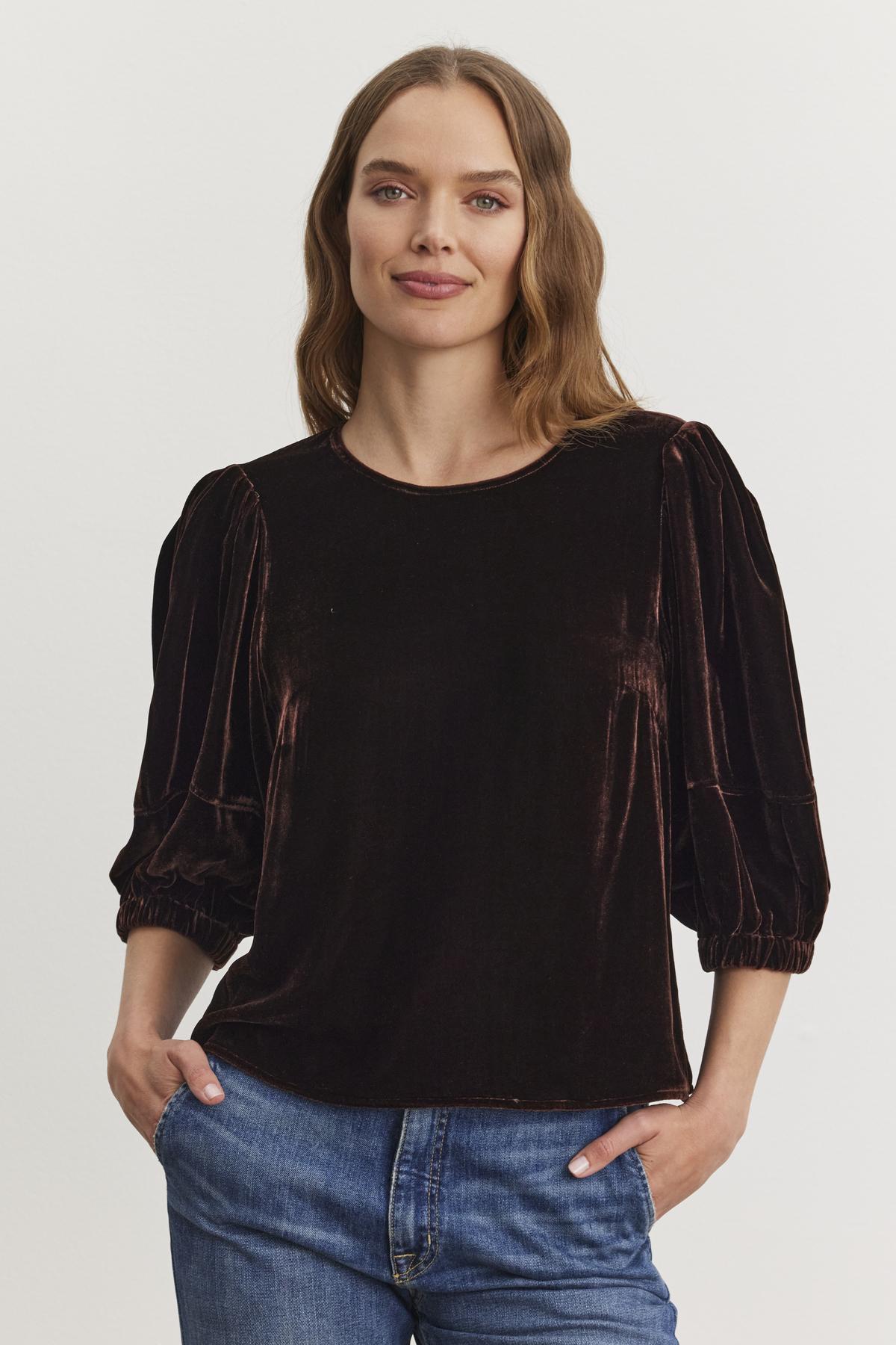 A woman with long hair wears the NANCY SILK VELVET TOP by Velvet by Graham & Spencer, paired with jeans, as she stands with her hands in her pockets against a plain white background.-38246590185665