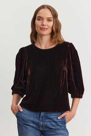 A woman with long hair wears the NANCY SILK VELVET TOP by Velvet by Graham & Spencer, paired with jeans, as she stands with her hands in her pockets against a plain white background.