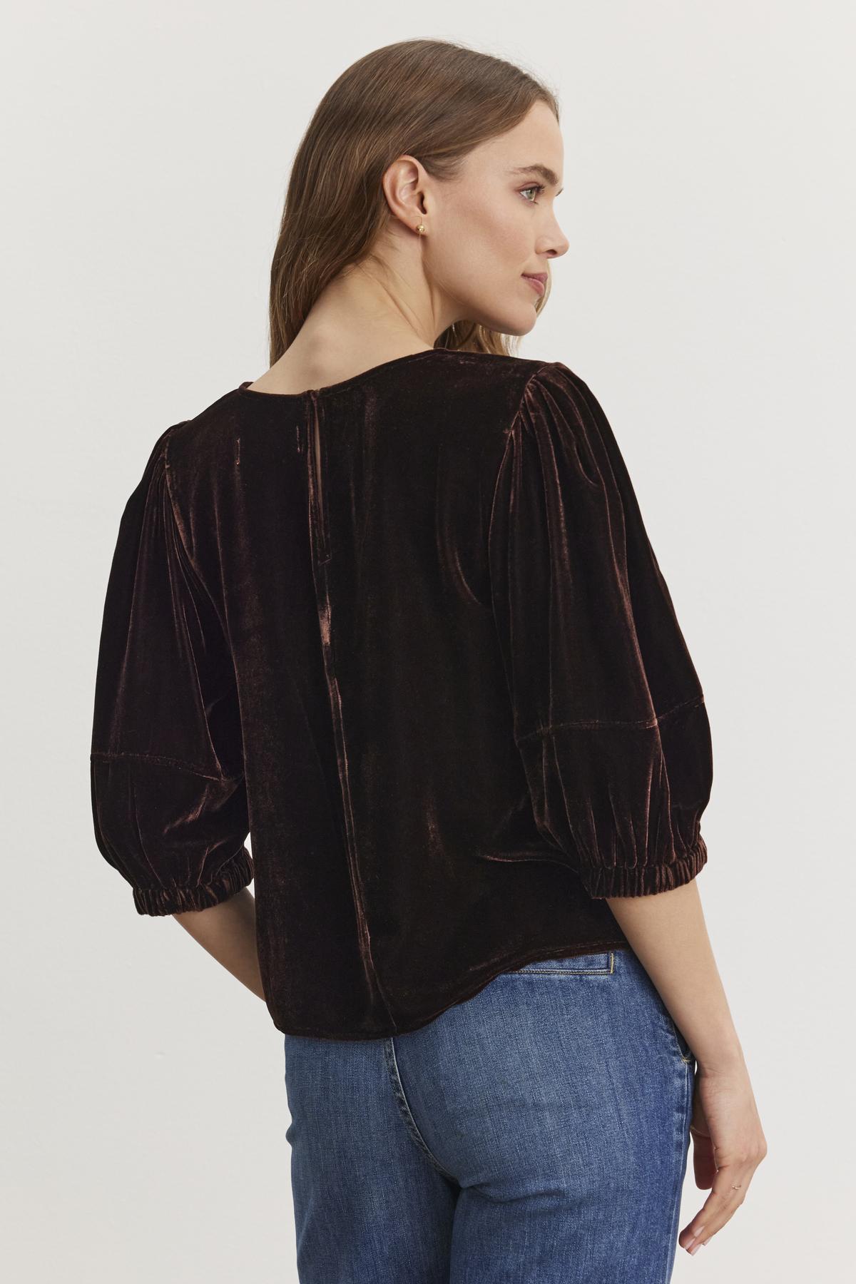 A woman wearing the NANCY SILK VELVET TOP by Velvet by Graham & Spencer, paired with blue jeans, stands with her back to the camera, looking to her right.-38246590251201