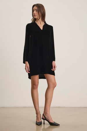 A woman stands exuding understated elegance in the SHERI SILK VELVET DRESS by Velvet by Graham & Spencer, paired with black high-heeled shoes against a plain background.