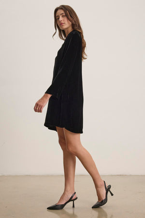 A woman in the SHERI SILK VELVET DRESS by Velvet by Graham & Spencer and black heels stands sideways, exuding understated elegance in a minimalistic setting.