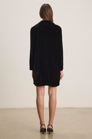 A person stands facing away on a plain background, wearing the SHERI SILK VELVET DRESS by Velvet by Graham & Spencer paired with black high heels, exuding an air of understated elegance.