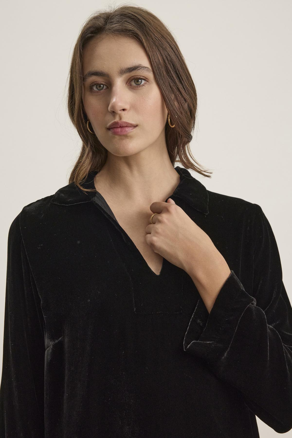 A woman with shoulder-length brown hair wears the SHERI SILK VELVET DRESS from Velvet by Graham & Spencer, featuring a refined style and a V-neckline, accompanied by gold hoop earrings, standing against a plain background.