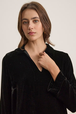 A woman with shoulder-length brown hair wears the SHERI SILK VELVET DRESS from Velvet by Graham & Spencer, featuring a refined style and a V-neckline, accompanied by gold hoop earrings, standing against a plain background.