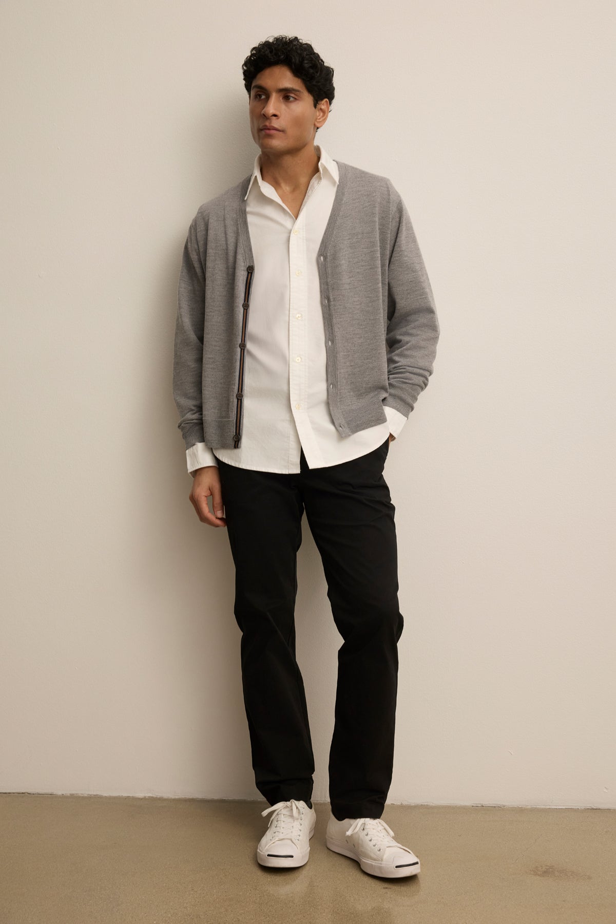 A man stands against a plain wall, wearing a white shirt, black pants, white sneakers, and the SILVIO CARDIGAN in gray by Velvet by Graham & Spencer.-39841094566081