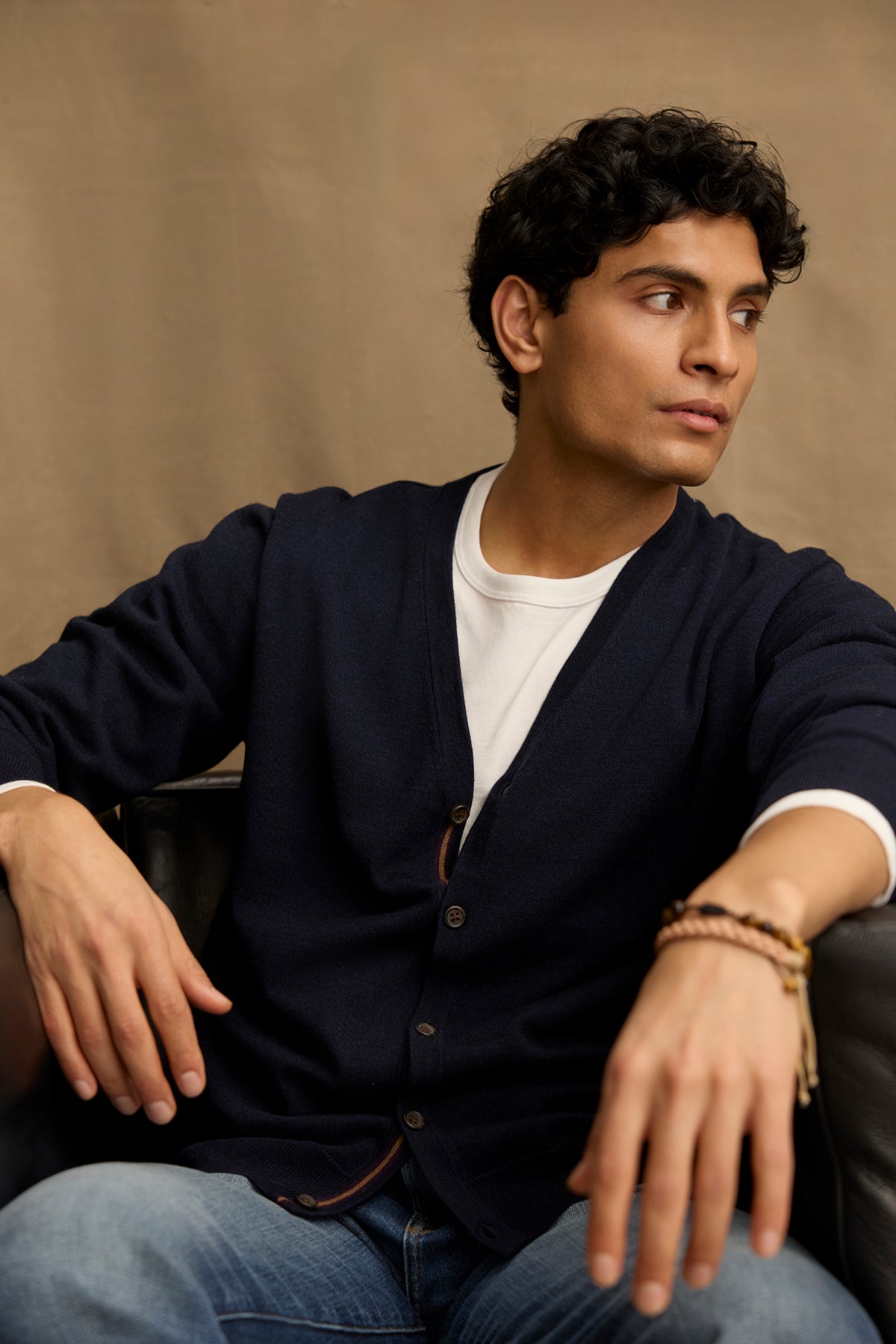 A man with curly dark hair sits on a black chair, wearing the SILVIO CARDIGAN by Velvet by Graham & Spencer—a navy V-neck with ribbed trims—over a white shirt and blue jeans, looking to his right with a neutral expression.-39543530750145