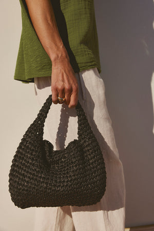 A close-up of a person holding a black Velvet by Graham & Spencer Slouch Bag, wearing a green top and white pants, with a focus on their hand and the bag.