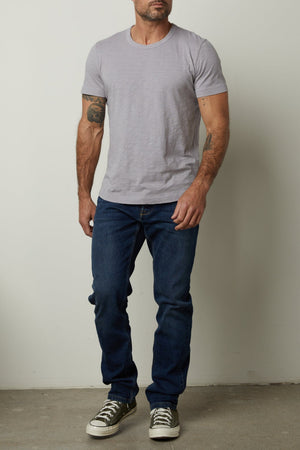 A man with short hair and tattoos on his arms stands against a plain wall, wearing a light gray, relaxed fit Velvet by Graham & Spencer AMARO TEE, blue jeans, and gray sneakers.