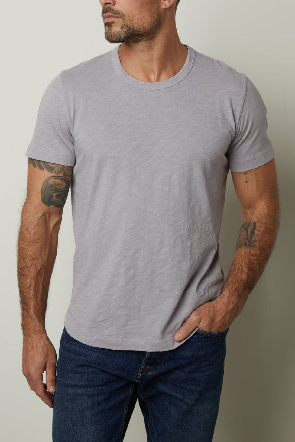 A person with short hair and a beard stands against a plain background, wearing a gray AMARO TEE by Velvet by Graham & Spencer and jeans, resting one hand in their pocket, and displaying tattoos on both arms.-37229768933569