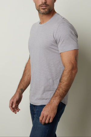 A person with short hair stands against a plain background, wearing a light gray Velvet by Graham & Spencer AMARO TEE and blue jeans, looking to the side.