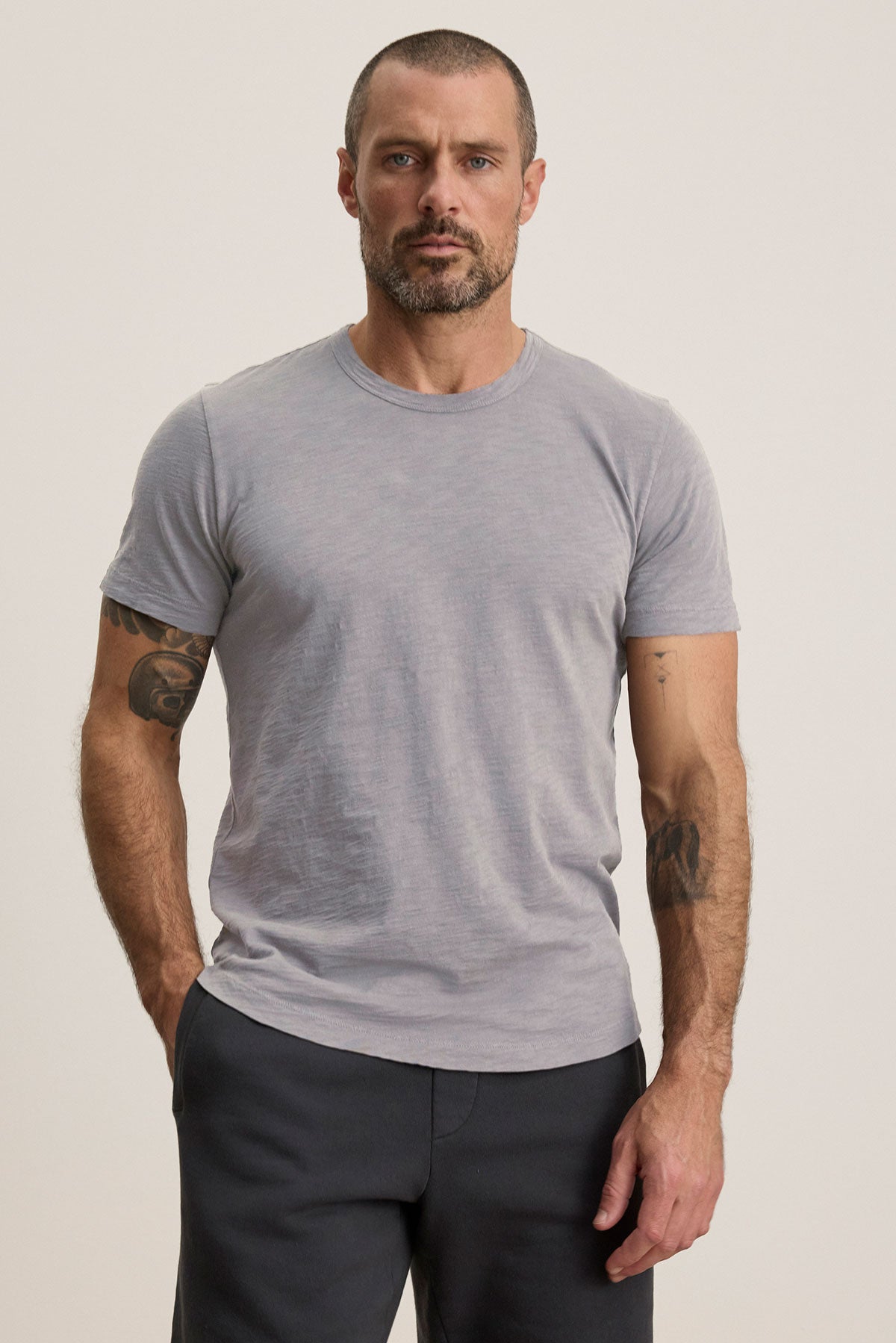 A man with short hair and a beard wears the AMARO TEE by Velvet by Graham & Spencer—a light gray Peruvian cotton crewneck t-shirt—paired with dark pants, standing with one hand in his pocket against a plain background.-40117510701249