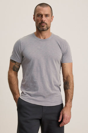 A man with short hair and a beard wears the AMARO TEE by Velvet by Graham & Spencer—a light gray Peruvian cotton crewneck t-shirt—paired with dark pants, standing with one hand in his pocket against a plain background.