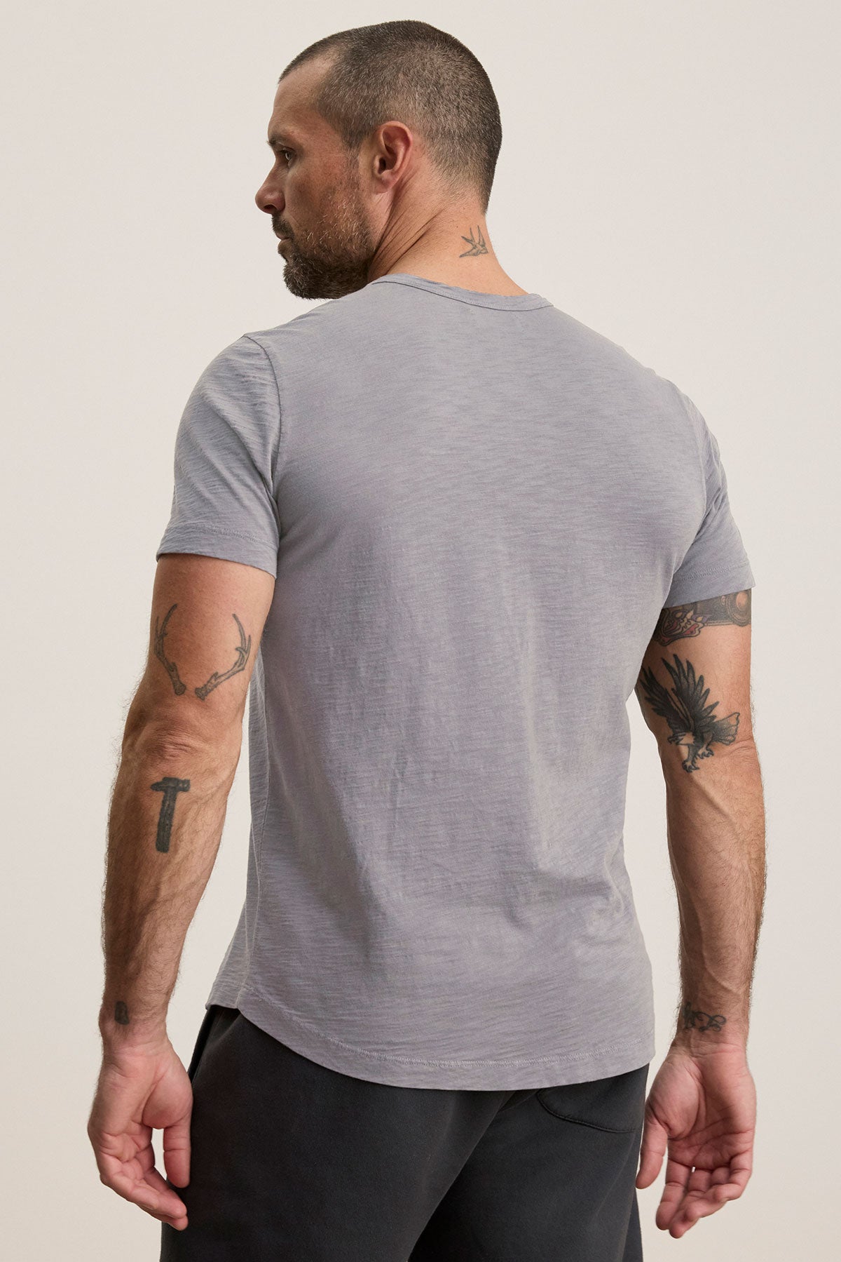 A man with short hair and tattoos stands with his back to the camera, wearing the light gray AMARO TEE by Velvet by Graham & Spencer and dark pants.