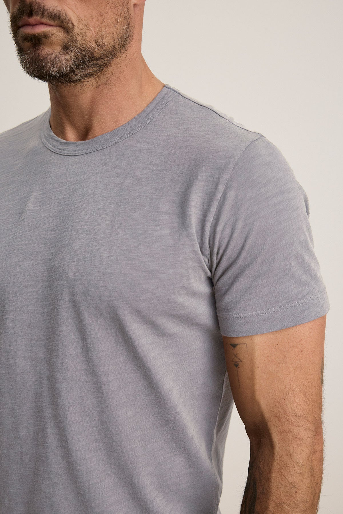 A man with facial hair is shown from shoulders to mid-torso, wearing the AMARO TEE by Velvet by Graham & Spencer—a light grey Peruvian cotton short-sleeve t-shirt with a classic crew neckline—against a plain background.-40117510766785