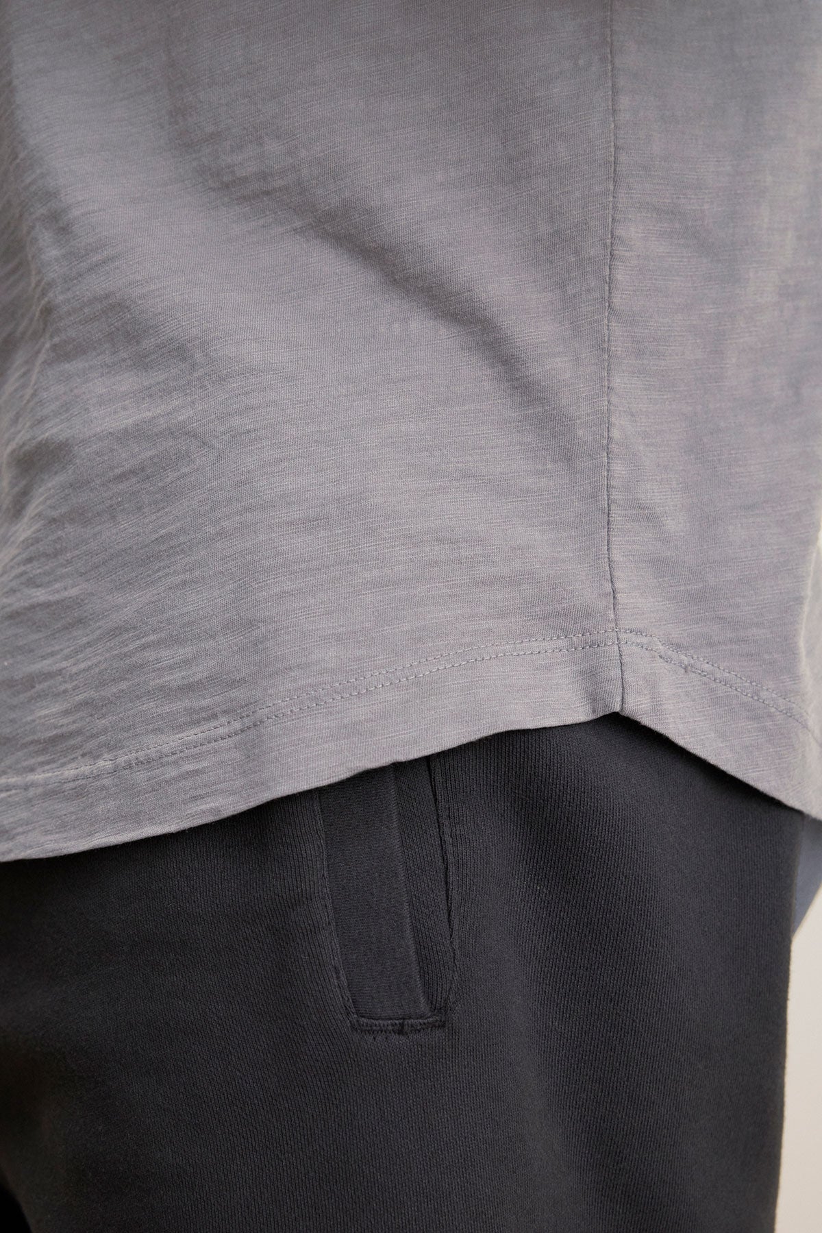 Close-up of the lower part of the light grey AMARO TEE by Velvet by Graham & Spencer and the upper part of dark pants, highlighting the tee's curved hemline and a side pocket on the pants.-40117510799553