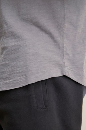 Close-up of the lower part of the light grey AMARO TEE by Velvet by Graham & Spencer and the upper part of dark pants, highlighting the tee's curved hemline and a side pocket on the pants.
