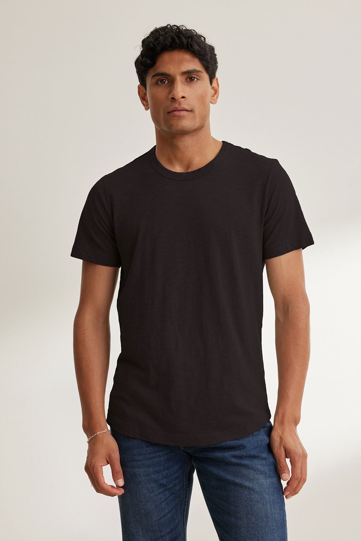 A dark-haired man stands against a light background wearing the AMARO TEE by Velvet by Graham & Spencer, made from soft Peruvian cotton. This plain black T-shirt features a classic crew neckline, perfect for layering. He's also wearing blue jeans.-39026123342017