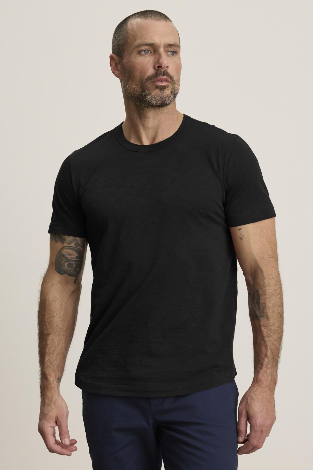 A man with short hair and a beard wears the AMARO TEE, a lightweight black Peruvian cotton T-shirt by Velvet by Graham & Spencer, paired with dark pants. Featuring a classic crew neckline, it’s perfect for layering as he faces slightly to the side against a plain background.