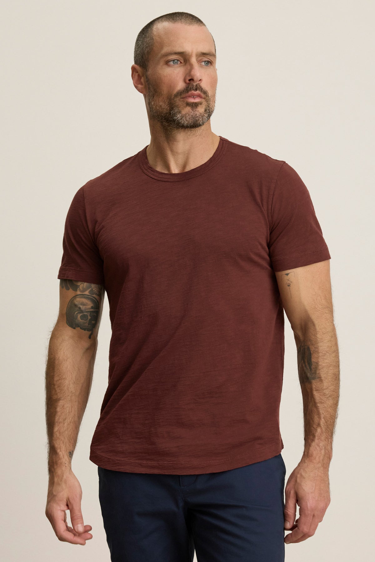 A man with short hair, a beard, and arm tattoos wears the maroon AMARO TEE by Velvet by Graham & Spencer—featuring Peruvian cotton and a classic crew neckline—paired with dark pants against a plain light background.
