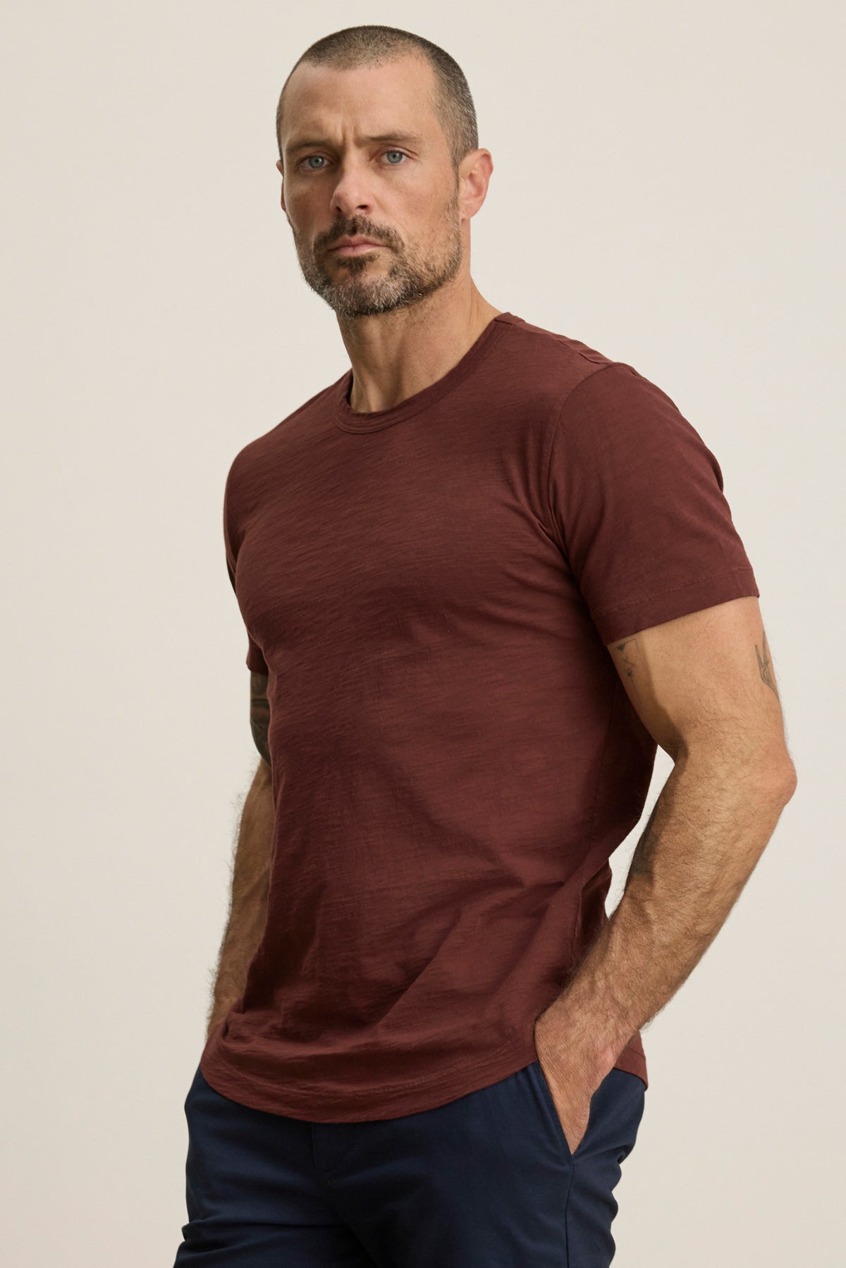 A man with a short beard and hair wears the AMARO TEE by Velvet by Graham & Spencer, a maroon Peruvian cotton crewneck, paired with navy pants, standing with hands in pockets against a plain background.-40054335701185