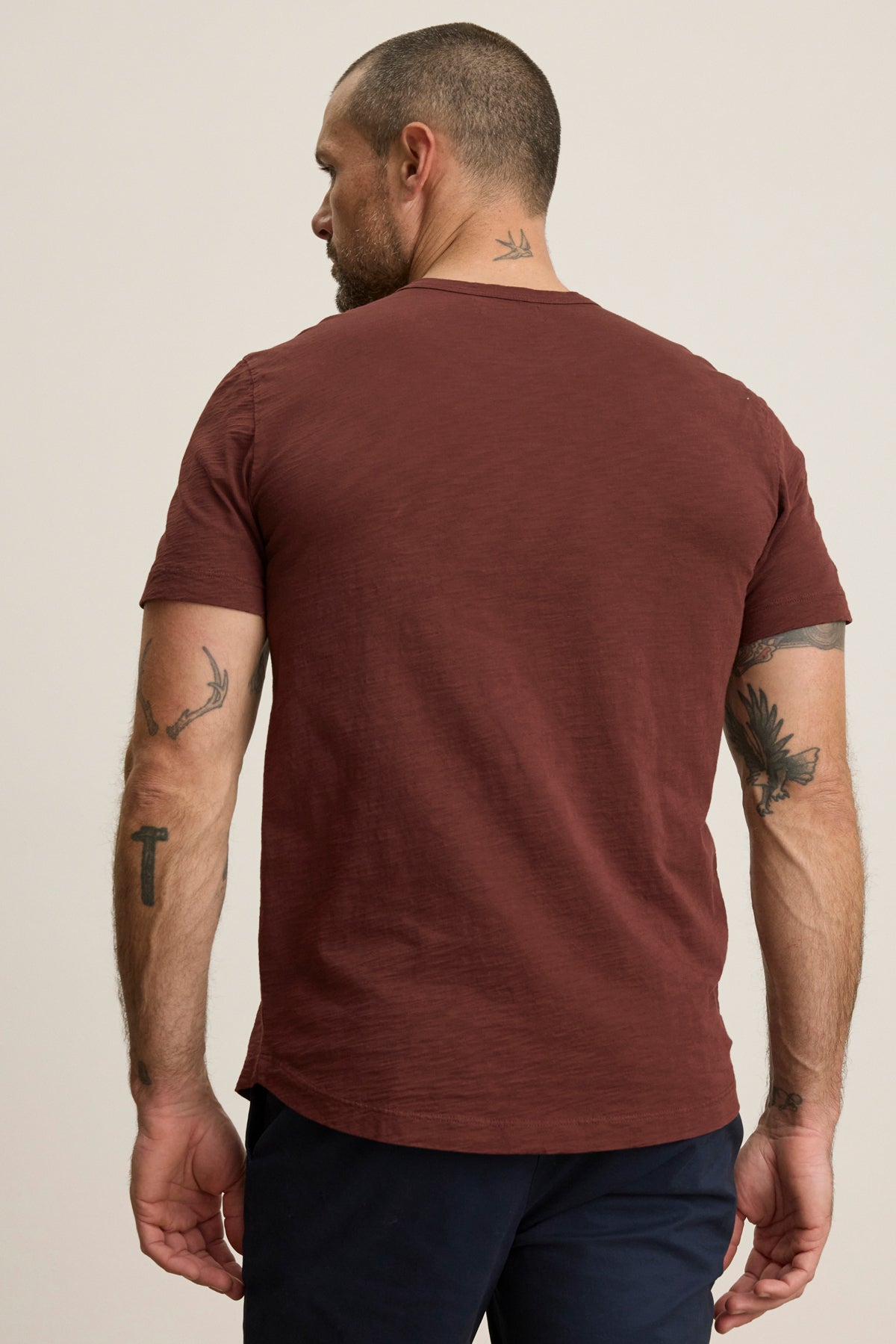 A man with short hair and tattoos wears the Velvet by Graham & Spencer AMARO TEE, a maroon Peruvian cotton crewneck, paired with dark pants. He stands with his back to the camera against a plain background.-40054335635649