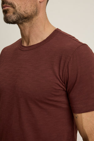 A man wears the AMARO TEE by Velvet by Graham & Spencer—a maroon short-sleeve crewneck—standing against a light background, shown from shoulders to mid-chest. This timeless tee is an ideal layering piece for any wardrobe.