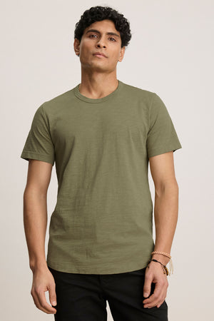 A person models the AMARO TEE by Velvet by Graham & Spencer—an olive green Peruvian cotton short-sleeve crewneck—paired with black pants, facing the camera against a plain light background.