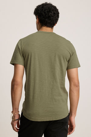 A man faces away from the camera, wearing the AMARO TEE by Velvet by Graham & Spencer—an olive green Peruvian cotton short-sleeve crewneck—paired with black pants against a plain light background.