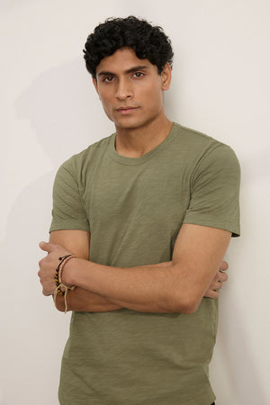 A person with short curly hair stands against a light-colored wall, arms crossed, wearing the AMARO TEE by Velvet by Graham & Spencer—an olive green Peruvian cotton t-shirt with a classic crew neckline.