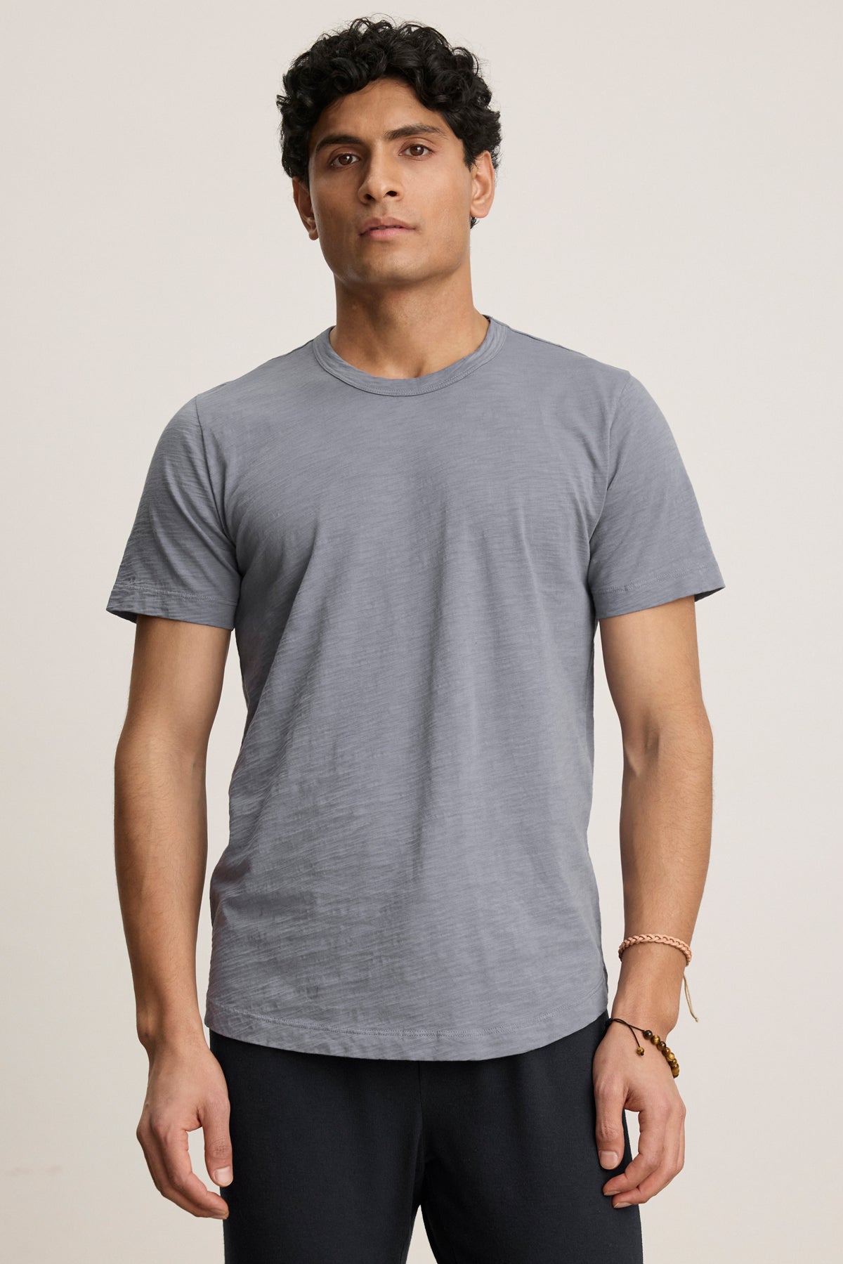 A man with short curly hair wears the AMARO TEE by Velvet by Graham & Spencer—an understated gray Peruvian cotton crewneck short-sleeve t-shirt—with dark pants, standing against a neutral background.-40054335537345