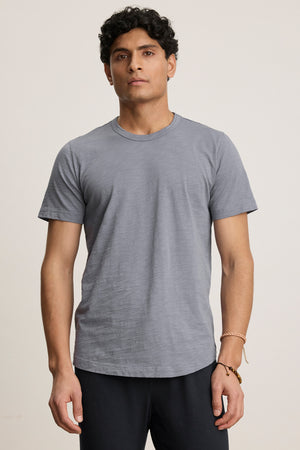 A man with short curly hair wears the AMARO TEE by Velvet by Graham & Spencer—an understated gray Peruvian cotton crewneck short-sleeve t-shirt—with dark pants, standing against a neutral background.