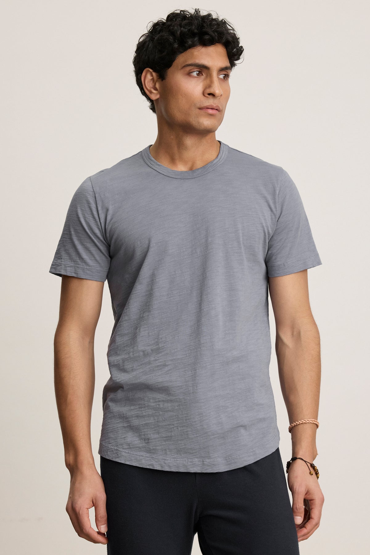 A man with short dark curly hair wears the AMARO TEE by Velvet by Graham & Spencer, a plain gray Peruvian cotton crewneck, and dark pants. He stands against a neutral background, looking slightly to his left.-40054335504577