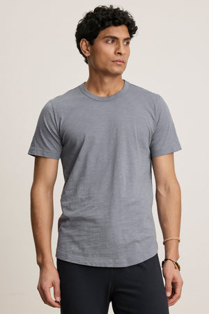 A man with short dark curly hair wears the AMARO TEE by Velvet by Graham & Spencer, a plain gray Peruvian cotton crewneck, and dark pants. He stands against a neutral background, looking slightly to his left.