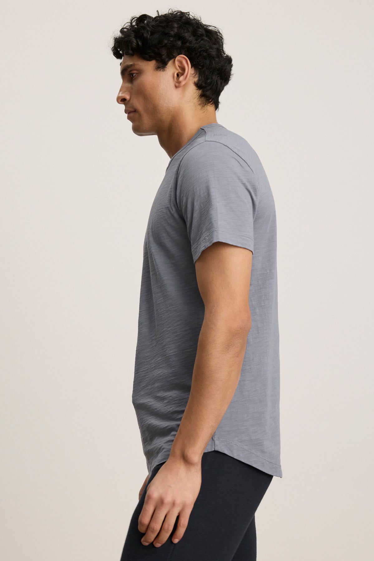 A person with short curly hair wears the AMARO TEE by Velvet by Graham & Spencer—a light gray Peruvian cotton t-shirt with a classic crew neckline—and dark pants, standing facing left against a plain background.-40054335471809