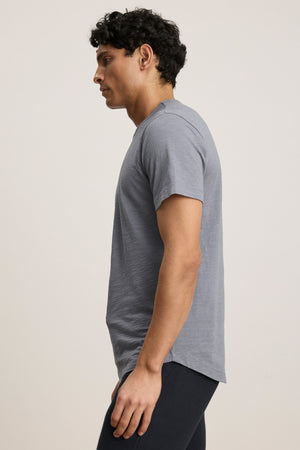 A person with short curly hair wears the AMARO TEE by Velvet by Graham & Spencer—a light gray Peruvian cotton t-shirt with a classic crew neckline—and dark pants, standing facing left against a plain background.