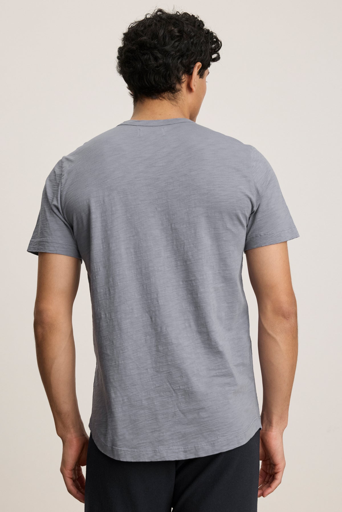 A person with short curly hair stands with their back to the camera, wearing a light gray AMARO TEE by Velvet by Graham & Spencer and dark pants, set against a plain background.-40054335439041