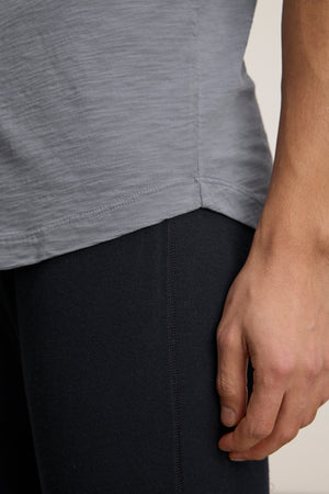 A close-up of a person's lower torso and hand, wearing the AMARO TEE by Velvet by Graham & Spencer in light gray Peruvian cotton and black pants—a perfect layering piece.