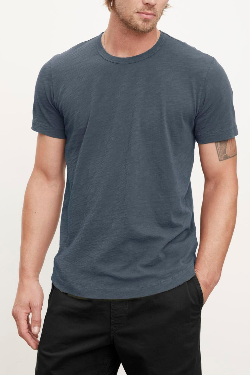A person with long hair wearing a relaxed stylish feel, casual, short-sleeved dark gray Velvet by Graham & Spencer AMARO TEE and black pants stands against a plain light background. They have a neutral expression and a tattoo on their left arm.