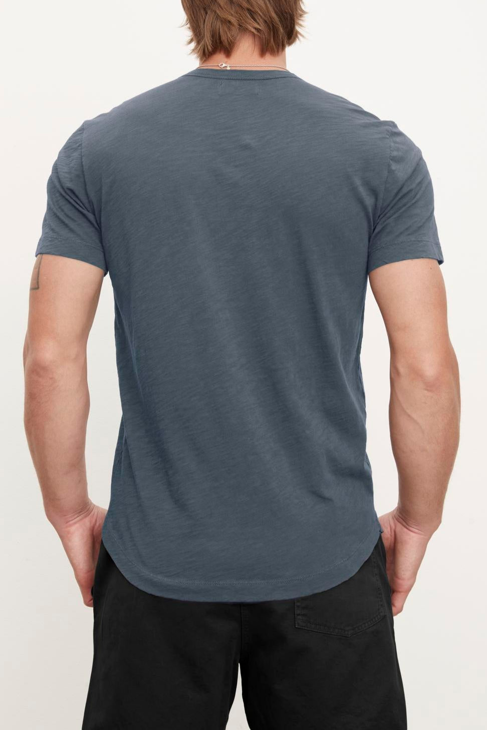 A person with shoulder-length hair is standing with their back to the camera, wearing a plain dark blue Velvet by Graham & Spencer AMARO TEE and black pants, exuding a relaxed stylish feel.-37386214441153